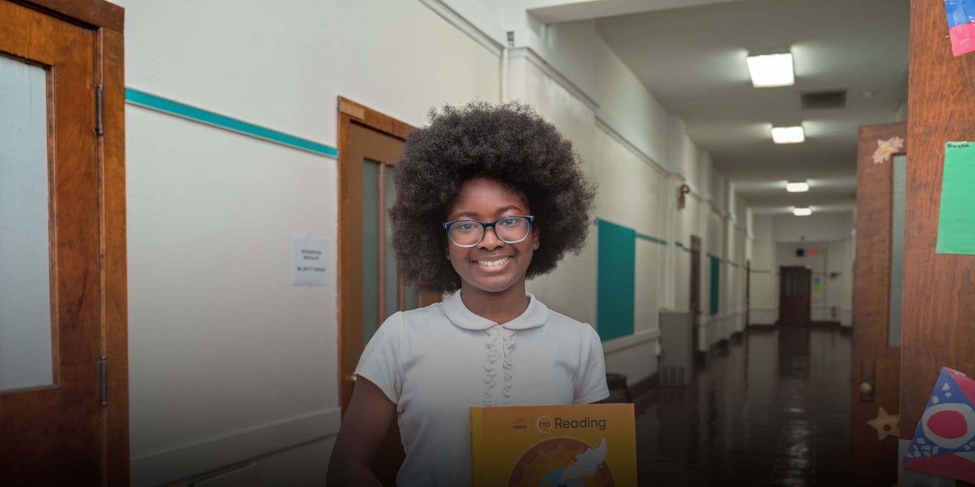 Smiling student in glasses holding a reading book in a school hallway.