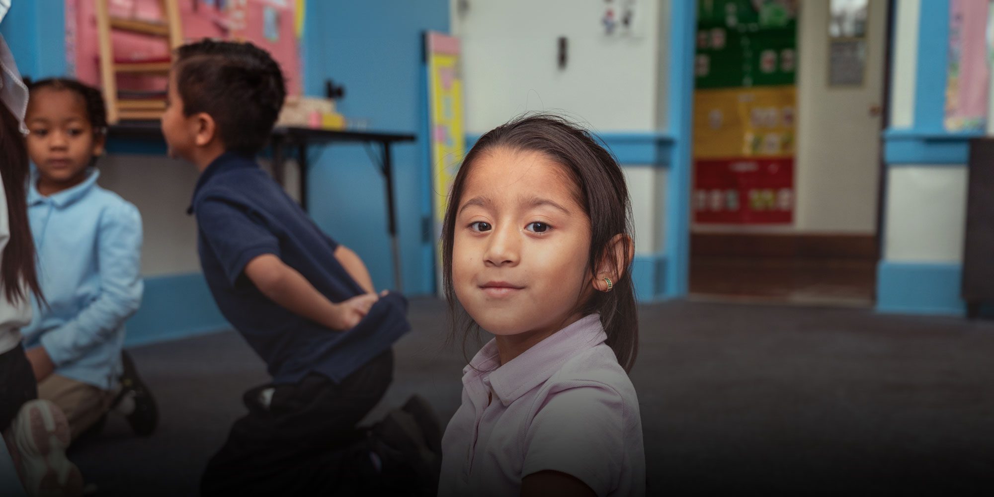 Young student in a pink uniform shirt looking at the camera while classmates play in the background.