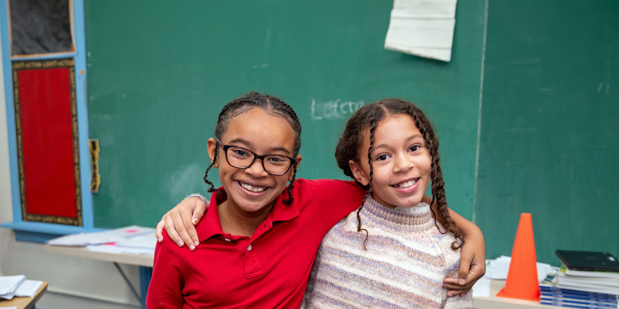 Two smiling students with arms around each other in a classroom.