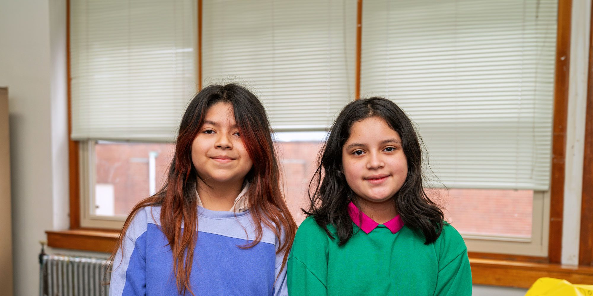 Two smiling students standing in front of classroom windows.